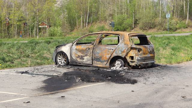 En svårt utbränd bil står övergiven på en parkeringsplats, omgiven av svartbrända bråte, med en gräsmatta och träd i bakgrunden.