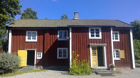 Ett rött tvåvåningshus i trä med vita lister, gula dörrar och ett tegeltak, beläget på en grusgård med gröna träd och blå himmel i bakgrunden.