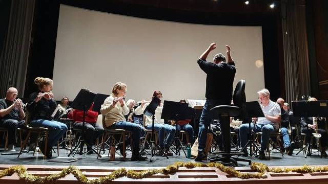 En dirigent leder en sittande orkester under en repetition på scenen; musiker spelar olika blåsinstrument med notställ framför sig.