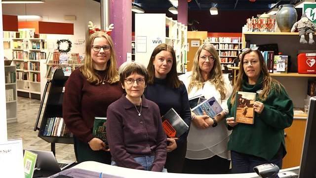 Fem kvinnor står tillsammans i ett bibliotek, var och en med en bok i handen, med bokhyllor och biblioteksutrustning i bakgrunden.