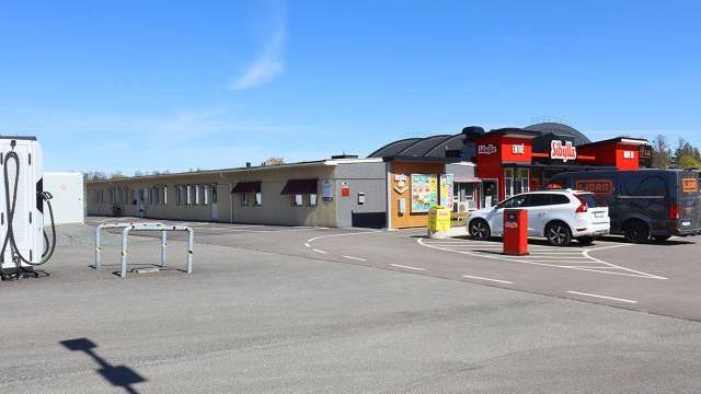 En bensinstation med närbutik och parkerade bilar en klar dag. Byggnaden har röda skyltar och stora fönster; en laddningsstation syns till vänster.