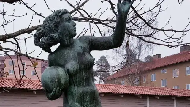 En bronsstaty av en kvinna som håller en boll och sträcker sig uppåt, placerad utomhus nära byggnader och lövlösa träd en molnig dag.