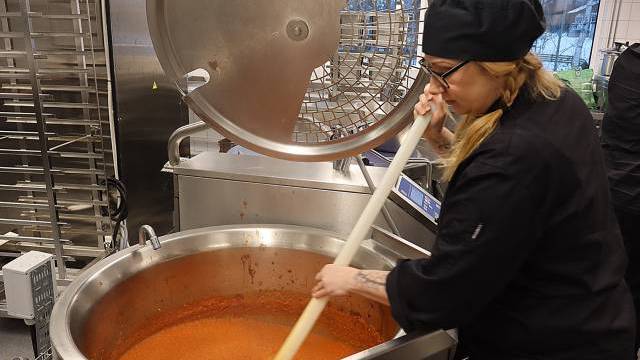 En kock i svart uniform och hatt rör om i en stor industriell gryta med tomatsås i ett storkök.