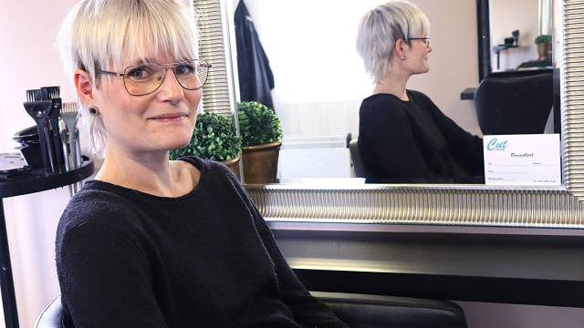 En kvinna med kort blont hår och glasögon sitter i en salongsstol vänd mot kameran; hennes spegelbild syns i spegeln bakom henne.