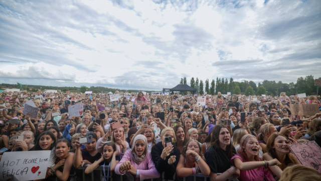 En stor skara entusiastiska fans, varav många håller upp skyltar och mobiltelefoner, står bakom en avspärrning vid en utomhuskonsert under en molnig himmel.