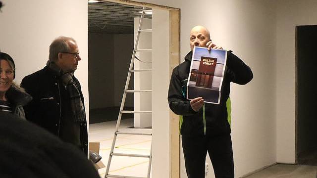 En man står i ett delvis färdigt rum och håller upp en affisch medan andra tittar på; en stege och byggmaterial syns i bakgrunden.