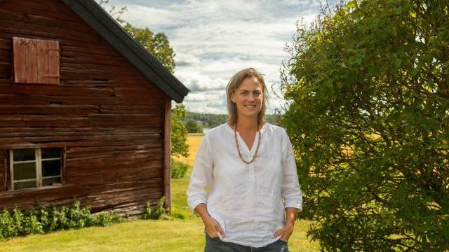 En kvinna i vit skjorta står vid en buske intill en väderbiten träbyggnad en solig dag, med åkrar i bakgrunden.