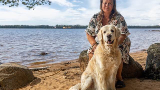 En kvinna i en blommig klänning sitter på en sten vid stranden, med en stor golden retriever som sitter framför henne på sandstranden.