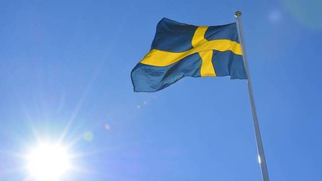 Den svenska flaggan med ett blått fält och ett gult nordiskt kors vajar på en flaggstång mot en klarblå himmel med solen skinande.
