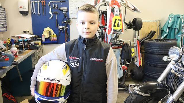 En pojke står i en verkstad och håller i en racinghjälm, med verktyg, däck och en gokart i bakgrunden.