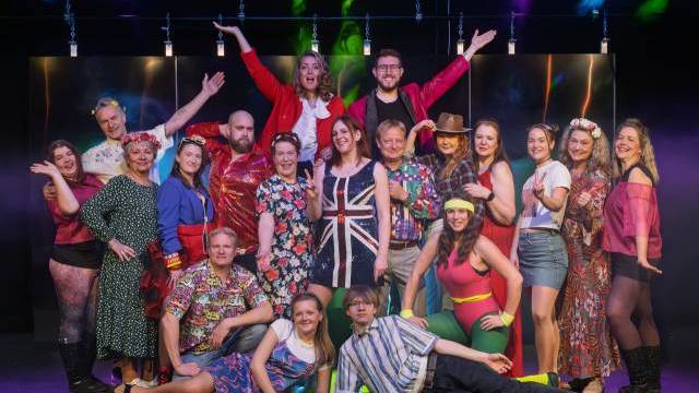 En grupp människor i färgglada retrokostymer poserar tillsammans på scenen i starkt ljus, några står, sitter och höjer armarna, framför en mörk bakgrund.