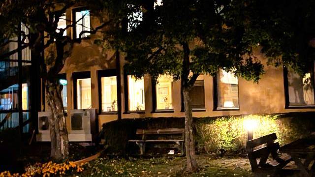 En svagt upplyst trädgård med bänkar och träd utanför en byggnad vars fönster lyser upp på natten.