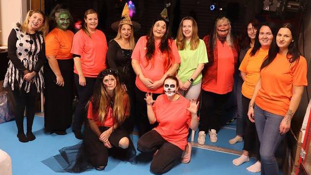 En grupp på tolv personer, några i orangea tröjor och andra i Halloweenkostymer, poserar tillsammans inomhus på ett blått golv.