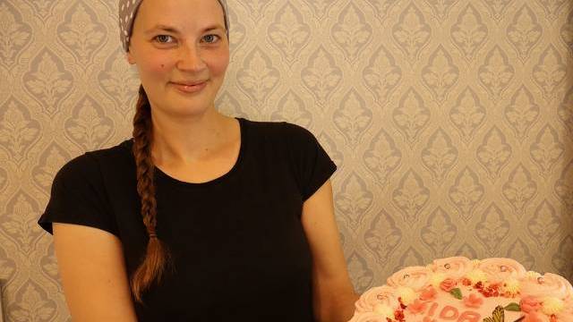 En kvinna med prickig huvudduk och svart skjorta håller en rosa frostad tårta dekorerad med namnet 
