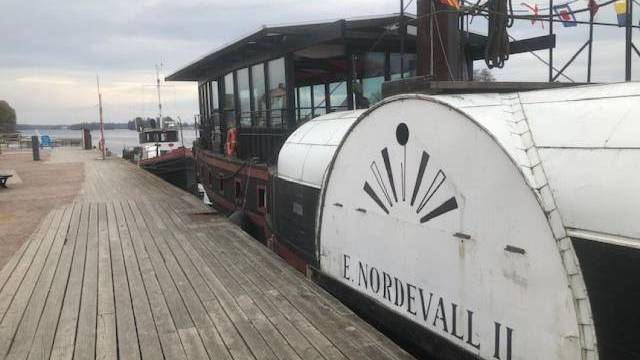 Ångbåten E. Nordevall II ligger vid en träbrygga med flaggor hängande ovanför och lugnt vatten i bakgrunden under en molnig himmel.