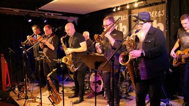 Ett band med sju musiker spelar brass- och stränginstrument på scenen, med notställ framför och en svart gardin och klubbskylt i bakgrunden.