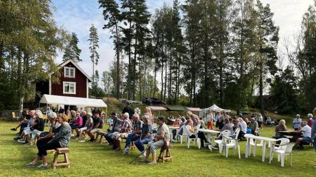 En grupp människor sitter på stolar och bänkar utomhus på en gräsplan nära ett rött hus och några tält, med träd i bakgrunden.