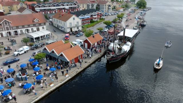 En stor folkmassa samlas längs en brygga vid vattnet med byggnader med röda tak, bredvid ett segelfartyg som ligger i hamn och en segelbåt på vattnet, i en småstadsmiljö.