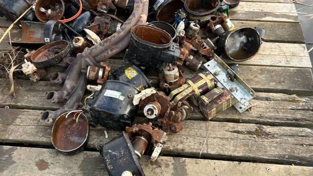 En hög med rostiga, trasiga rördelar och ventiler ligger på en träbrygga intill ett vattendrag.