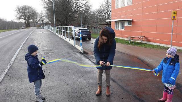 En kvinna klipper av ett blågult band som hålls av två barn vid en vägkant intill en byggnad för att markera en invigning.
