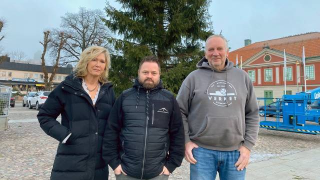 Tre vuxna står utomhus framför ett stort vintergrönt träd och byggnader, iklädda vinterjackor och fritidskläder.