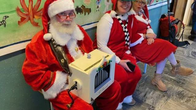 Tre personer i festliga dräkter, varav en är utklädd till tomte och håller i en vit låda, sitter på en bänk i en dekorerad hall med en väggmålning bakom sig.