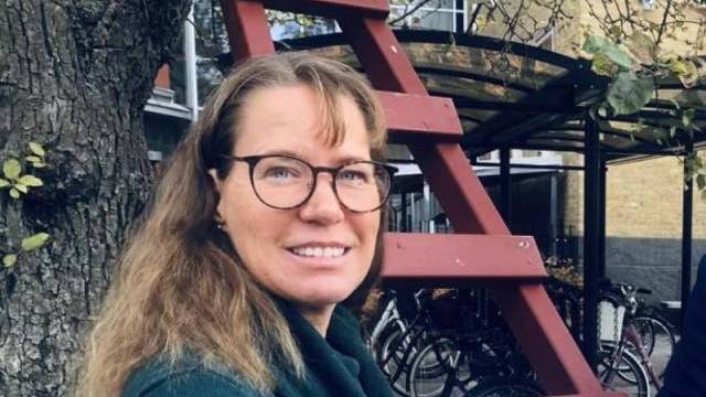 En person med glasögon och långt hår står utomhus nära ett träd och en röd träkonstruktion och håller en broschyr i handen. I bakgrunden syns cyklar och en del av en byggnad.