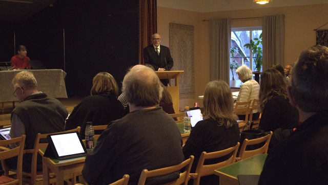 En man står på ett podium och talar till en publik som sitter vid bord med bärbara datorer och anteckningsböcker i ett konferensrum.