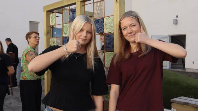 Två unga kvinnor står utomhus framför en kakelutställning och gör båda tummen upp-tecknet med höger hand nära hakan.