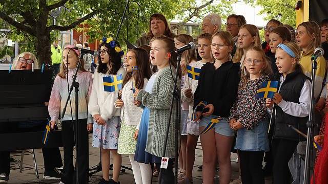 En barnkör sjunger utomhus på en scen och håller i små svenska flaggor. Bakom dem står flera vuxna och i bakgrunden syns träd och byggnader.