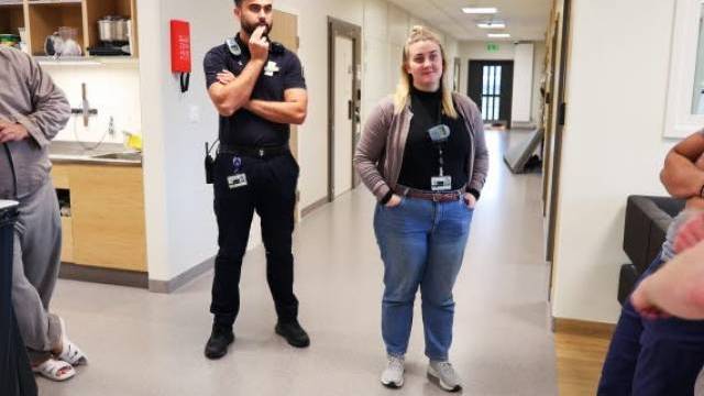 Två personer med arbetsmärken står i en korridor, den ena med armarna i kors och den andra med händerna i fickorna. Andra personer är delvis synliga i närheten.