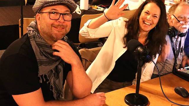 Tre vuxna sitter vid ett bord med mikrofoner och ljudutrustning; en kvinna vinkar och ler mot kameran medan de andra tittar på.