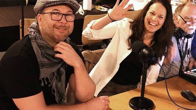 Tre vuxna sitter vid ett bord med mikrofoner och ljudutrustning; en kvinna vinkar och ler, medan de andra tittar mot kameran.