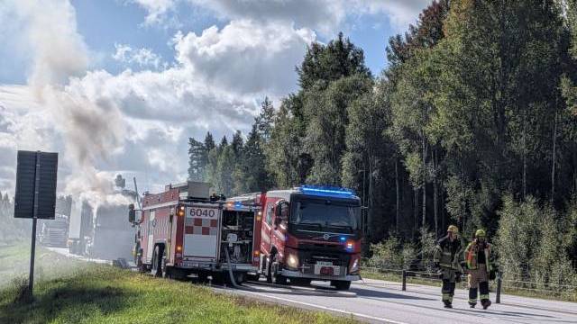 Två brandbilar med blinkande ljus står parkerade på en väg nära träden, medan rök stiger upp i bakgrunden och två brandmän går längs vägkanten.