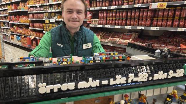 En butiksanställd står bakom en LEGO-modell av en stormarknad, där öppettiderna anges på svenska, med hyllor fyllda med varor i bakgrunden.