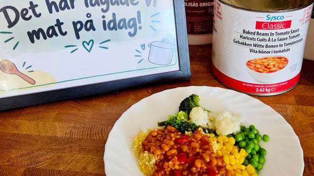 En tallrik med bakade bönor, majs, ärtor, broccoli och blomkål står bredvid en burk bönor och en skylt på svenska där det står: ”Det här lagade vi mat på idag!”.
