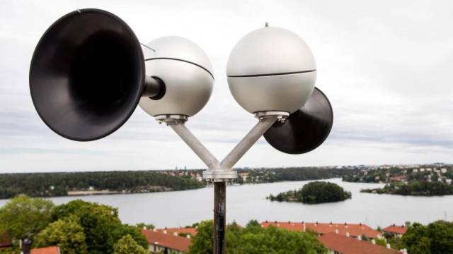 Ett kluster av utomhushögtalare monterade på en metallstolpe har utsikt över ett vattendrag och omgivande byggnader och träd.