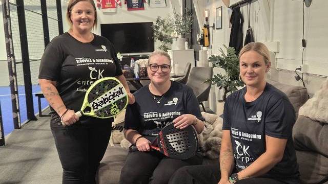 Tre kvinnor i svarta tröjor sitter och står inomhus med padelracketar i händerna, medan sportutrustning och tröjor syns i bakgrunden.