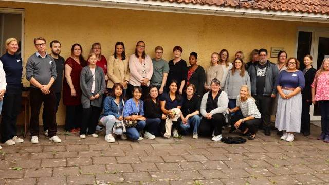 En grupp vuxna poserar tillsammans utomhus framför en gul byggnad, stående och sittande på en asfalterad yta.