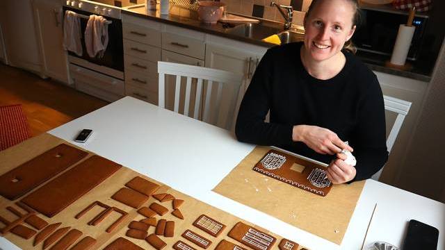 En kvinna sitter vid ett köksbord och sätter ihop delar till ett pepparkakshus, med glasyr i handen och olika dekorerade pepparkaksplattor framför sig.