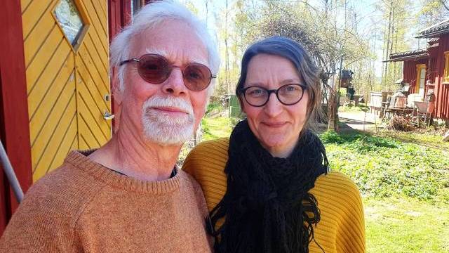 En äldre man och en kvinna står tätt intill varandra utomhus vid en röd träbyggnad och ler mot kameran en solig dag.