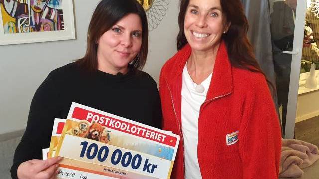 Två kvinnor står inomhus och ler; den ena håller stora lottericheckar, bland annat en på 100.000 kr, med 