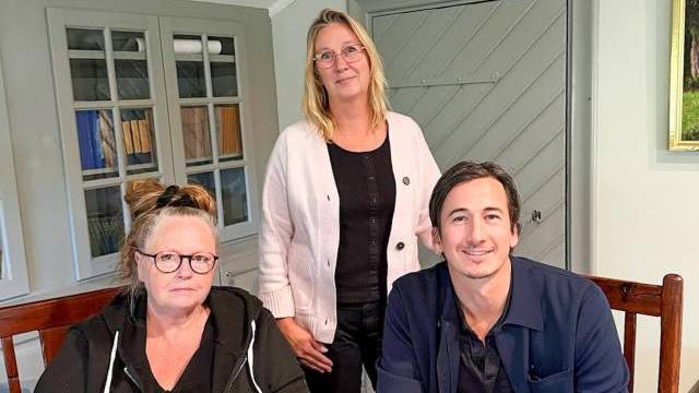 Tre vuxna inomhus, två sittande vid ett bord och en stående bakom dem, alla klädda i vardagskläder och tittar in i kameran.