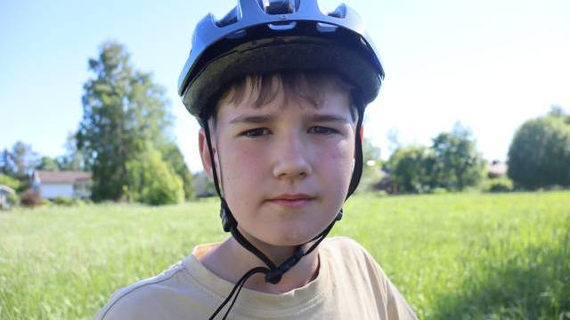 En pojke med cykelhjälm står utomhus på en gräsplätt en solig dag och tittar rakt in i kameran.
