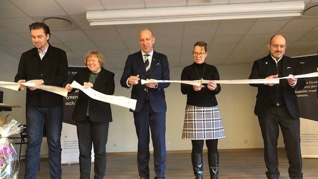 Fem personer står inomhus och håller i ett vitt band under en klippningsceremoni. Fyra av dem håller i saxar och förbereder sig för att klippa bandet.