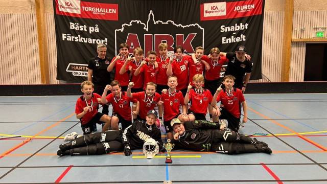 Ett ungdomsinnebandylag i röda tröjor poserar inomhus med medaljer och pokal framför en stor AIK-banderoll.