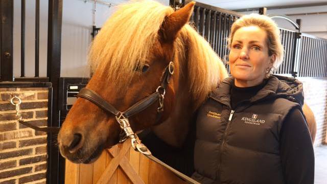 En kvinna står bredvid en brun häst i ett stall och håller i dess lina. Hästen har en tjock, ljus man och är vänd framåt.