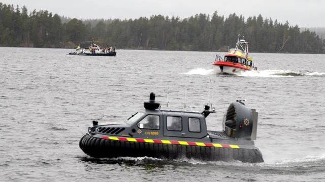 En svart och gul svävare färdas på en sjö, och i bakgrunden syns två räddningsbåtar nära en skogskantad strand.