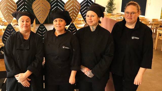 Fyra kvinnor i svarta kockuniformer står sida vid sida i en matplats med bord och dekorativa bladformade paneler i bakgrunden.