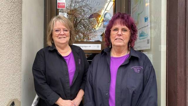 Två kvinnor med glasögon och matchande lila skjortor med svarta jackor står framför en butiksentré och ler mot kameran.